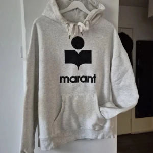 Grå hoodie från Marant med logga - Säljer en ljusgrå hoodie från Marant med svart logga och text framtill. Tröjan har huva med snörning, stor känguruficka och långa ärmar. Ingen lapp i nacke.Perfekt för dig som gillar en avslappnad och stilren look.