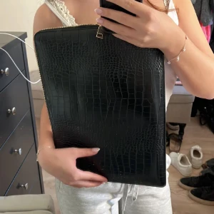 Svart datorväska i croco-imitation - Säljer en stilren svart datorväska med croco-mönstrad yta och guldfärgad dragkedja. Väskan är rektangulär och har plats för laptop eller dokument. Perfekt för dig som vill ha en clean och trendig look till plugget eller jobbet.