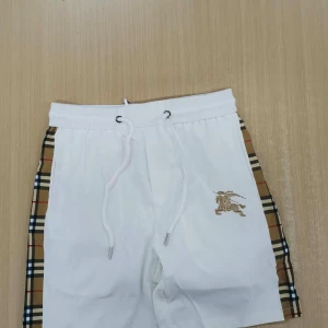 Vita Burberry shorts med rutiga sidor - Snygga vita shorts från Burberry med klassiska beige, svarta och röda rutiga paneler på sidorna. Shortsen har snörning i midjan och ett guldfärgat broderat riddaremblem på ena benet. Perfekta för en trendig och avslappnad look.