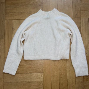 Vit stickad tröja från & Other Stories - Supermjuk och ribbstickad vit tröja från & Other Stories Los Angeles Atelier. Tröjan har hög hals, lång ärm och en lite boxig, croppad passform. Materialmix med alpacka, ull och nylon gör den extra mysig och perfekt för kyliga dagar.