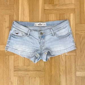 Ljusblå jeansshorts från Hollister - Snygga ljusblå jeansshorts från Hollister med råa, fransiga kanter och klassiska fem fickor. Shortsen har låg midja, slitna detaljer och gulddetaljer på bakfickorna. Perfekta för varma sommardagar och en avslappnad look.