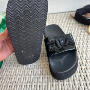 Svarta sandaler från Fila - Svarta sandaler från Fila med glansig rem och broderad logga framtill. Platt sula och öppen tå ger en chill vibe. Perfekta för sommardagar eller att glida runt i hemma. Snygg och enkel design som passar till det mesta.