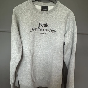 Grå sweatshirt från Peak Performance strl 160 - Snygg grå sweatshirt från Peak Performance med svart broderad logga på bröstet. Klassisk rund halsringning och långa ärmar. Mjuk och skön bomullsblandning, perfekt för chill eller skola. Enkel stil som funkar till allt.