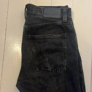 Svarta jeans från Nudie Jeans, W31 L30 - Snygga svarta jeans från Nudie . Jeansen har silverfärgade och kopparfärgade nitar, samt Nudie Jeans-logga på bakfickan. Perfekta för dig som gillar en stilren och tidlös look. Aldrig använda
