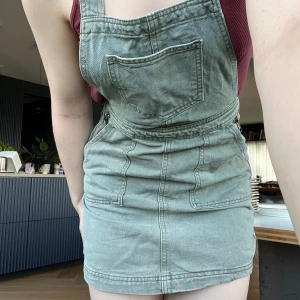 Grön jeansklänning med hängslen - Snygg kort grön jeansklänning med hängslen och fickor både fram och bak. Klänningen har klassisk förklädesstil med raka axelband och dragkedja på sidan. Perfekt att styla med en t-shirt eller linne under för en chill look.