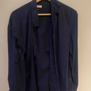 Mörkblå skjorta V3rsace - En stilren mörkblå skjorta från V3rsace med klassisk krage och knäppning framtill. Skjortan har lång ärm och är tillverkad i ett mjukt bomullstyg som känns skönt mot huden. Perfekt för dig som gillar en enkel och clean look.