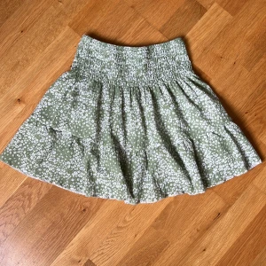 Grön blommig kjol från Gina Tricot - Supersöt kort kjol från Gina Tricot i ljusgrönt med vitt blommönster. Kjolen har smock i midjan, volanger och är gjord i ett lätt och luftigt tyg. Perfekt för dig som gillar en feminin och trendig stil.
