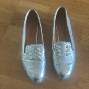 Silvriga loafers med pärldetaljer - Glansiga silvriga loafers med dekorativa pärlor framtill och broguemönster. Skorna har spetsig tå och platt sula, samt små nitar längs sulkanten. Perfekta för dig som vill sticka ut med en unik och trendig look.