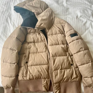 Beige pufferjacka med huva - Snygg beige pufferjacka med huva och ribbade muddar. Jackan har dragkedja framtill, två fickor med knapp och quiltad design. Perfekt för kyliga dagar och ger en clean look till din outfit.