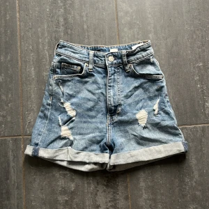 Blåa mom jeansshorts med slitningar - Snygga blå mom shorts från &Denim med ultrahög midja och uppvikta benslut. Shortsen har slitna detaljer framtill och bakfickor, klassisk femficksmodell och är gjorda i jeansmaterial. 