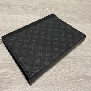 Louis Vuitton handväska  - Snygg svart kuvertväska  med klassiskt monogrammönster. Väskan är tillverkad i skinn och har en silverfärgad dragkedja upptill. Perfekt för att bära det viktigaste och ger en lyxig touch till din outfit.