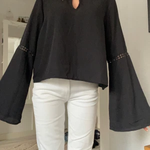 Svart blus med vida ärmar - Säljer en svart blus med vida, långa ärmar och snygg detalj med spetsinslag vid armbågen. Blusen har en liten v-ringning framtill och är i ett lätt, flowy material som ger en cool och avslappnad vibe.