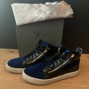 Blåa sneakers från Giuseppe Zanotti - Snygga sneakers i blå sammet och svart lack från Giuseppe Zanotti. Skorna har guldfärgade dragkedjor på sidorna, snörning och vit platt sula. Ikonisk logga i guld på plösen ger en lyxig vibe. Perfekta för dig som vill sticka ut med stil.