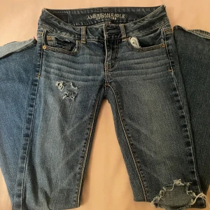Blå slitna jeans från American Eagle - Snygga blå jeans från American Eagle med slitningar på låret och vid benslutet. Klassisk femficksmodell med broderade bakfickor och låg midja. Jeansen har en rak passform och är tillverkade i mjukt denimtyg för en bekväm känsla.