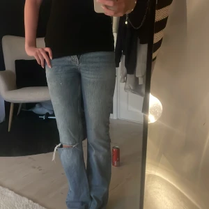 Blå bootcut jeans med hål - Säljer ett par blå bootcut jeans med snygg slitning och hål på ena knät. Säljer då de blivit för små i midjan, midwaist. Jag är 170 och de går ner till marken 