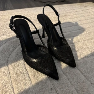 Svarta spetsiga pumps med spetsdetalj - Snygga svarta pumps med spetsig tå och klack. Skorna har tunna remmar med spänne runt hälen och ovandelen är täckt av elegant spetsmaterial. Perfekta för dig som vill ha en chic och feminin look med extra edge.