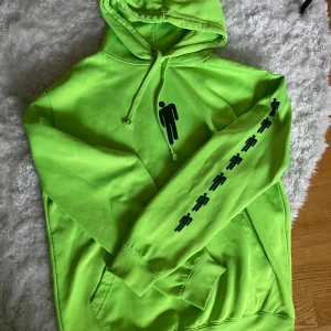 Billie Eilish Hoodie, neongrön - Säljer en neongrön Billie Eilish hoodie med svart figurmönster på bröstet och längs ena ärmen. Tröjan har huva med snörning och en stor magficka. Tröjan är oanvänd, köpt från Billie Eilish Merch
