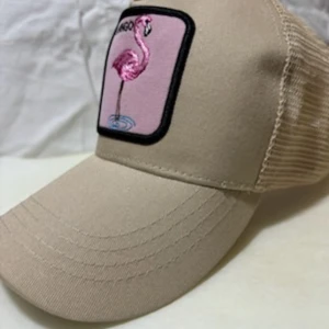 Beige truckerkeps med flamingo-patch - Snygg beige truckerkeps med mesh baktill och justerbar snapback. Framtill sitter en rosa patch med broderad flamingo och svart kant. Perfekt accessoar för dig som vill sticka ut med en cool detalj.