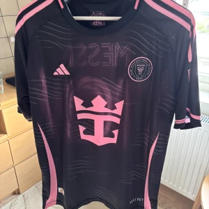 Inter Miami Messi 10 matchtröja Adidas - Snygg Inter Miami fotbollströja från Adidas med Messi 10-tryck på ryggen. Tröjan är svart med rosa detaljer, klubbmärke och stora grafiska tryck framtill. Tillverkad i lätt och ventilerande material, perfekt för fotbollsplanen eller att bära som supporter.
