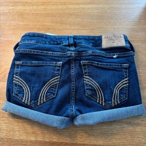 Mörkblå jeansshorts från Hollister - Snygga mörkblå jeansshorts från Hollister med klassisk femficksdesign och uppvikta benslut. Shortsen har kontrastsömmar och en läderpatch med logga bak. Perfekta för varma dagar och enkel att matcha med allt.