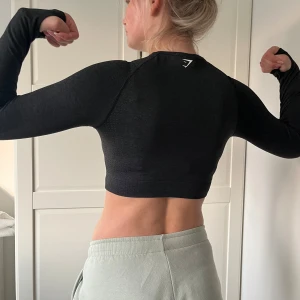Svart croppad träningströja Gymshark - Säljer en svart croppad långärmad träningströja från Gymshark. Tröjan har tight passform, rund halsringning och tumhål vid ärmsluten. Perfekt för gymmet eller löprundan, med diskret logga på bröstet.