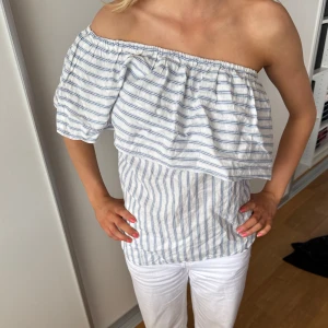 Randig offshoulder topp i bomull - Säljer en vit och ljusblå randig offshoulder topp med volangdetalj över bröstet. Toppen har en oneshoulder-design och är tillverkad i tunn bomull, perfekt för varma dagar. Snygg och luftig med avslappnad passform, passar till både jeans och shorts. ☺️Köpte här på plick men tröjan var för stor för mig så säljer därför vidare!❤️ Tröjan har inga defekter och är i fint skick🙏Vet inte storleken på tröjan men jag tycker den sitter som storlek M