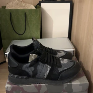 Valentino Garavani Rockrunner sneakers - Snygga Valentino Garavani Rockrunner sneakers i svart och grått med coolt kamouflagemönster. Skorna har mesh och mockadetaljer, svarta snören och ikoniska nitar på sulan. Perfekta för dig som vill sticka ut med stilrena och exklusiva sneakers.