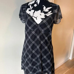Svart rutig meshklänning med rosett - Supersöt kort svart klänning i mesh med vitt rutmönster, liten rosett vid urringningen och v-ringad hals. Korta, transparenta ärmar med vågig kant ger en extra detalj. Använd 2 gånger men för liten för mig skulle mer säga M
