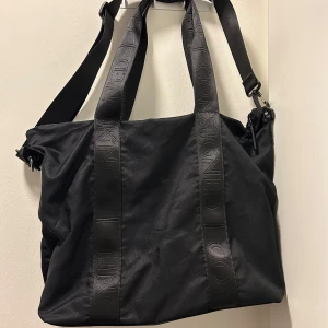 Svart axelväska från Consigned - Stor svart axelväska från Consigned med breda axelband och detaljer i samma färg. Väskan har diskret logga på banden och dragkedja upptill. Perfekt för att få plats med allt du behöver till skolan eller stan.