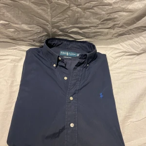 Ralph Lauren skjorta  - Klassisk mörkblå skjorta från Ralph Lauren i custom fit. Skjortan har button down-krage, vita knappar och den ikoniska broderade loggan på bröstet. Storlek M, hör av er vid frågor perfekt nu för sommaren 