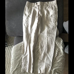 Beiga linnebyxor från Bläck, stl 46 - Snygga beiga linnebyxor från Bläck i tailored fit. Byxorna har klassisk kostymskärning med hällor för bälte, sidfickor och bakfickor med knapp. Perfekta för en fräsch och stilren look under varmare dagar.