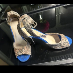 Leopardmönstrade pumps med blå sula - Snygga pumps med leopardmönster och öppen tå. Skorna har en blå innersula och smal klack, samt justerbart spänne runt ankeln. Perfekta för dig som vill sticka ut med djurmönster och coola detaljer.