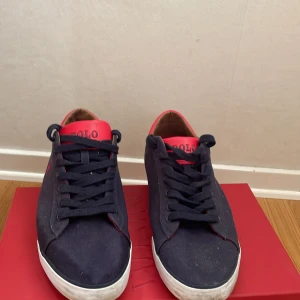 Mörkblå sneakers från Polo Ralph Lauren - Snygga mörkblå sneakers från Polo Ralph Lauren med röda detaljer och logga på sidan. Skorna är ett väldigt bra skick som jag skulle säga är 9/10 och är bara använda nån enstaka gång. Skorna har snörning, vit sula med röda och blå ränder samt lågt skaft. Ovandelen är i textil och insidan är ljusbrun. Perfekta för en stilren och sportig look.