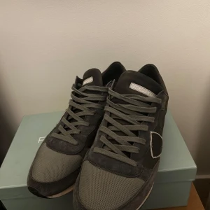 Grå sneakers från Philippe Model - Snygga grå sneakers från Philippe Model med coola detaljer i mocka och mesh. Skorna har en vit och svart randig sula, snörning och den klassiska skölden på sidan. Perfekta för dig som gillar stilrena 