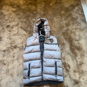 Grå dunväst från Philipp Plein - Snygg grå dunväst från Philipp Plein med stor huva och logotyp på bröstet. Västen har svarta dragkedjor på fickorna och längs framsidan samt tydlig branding på huvkanten. Perfekt för lager-på-lager och streetstyle.