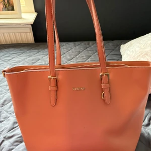 Korallrosa axelväska från Vabene - Snygg axelväska i korallrosa från Vabene med stilrena guldiga detaljer och justerbara axelremmar. Väskan har flera fack inuti, dragkedja upptill och är tillverkad i slätt syntetmaterial. Perfekt för dig som vill ha plats för allt du behöver under dagen.