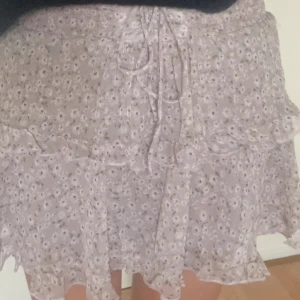 Ljusblommig volangkjol med snörning - Supersöt kort kjol i ljuslila med små blommor, volangkant och snörning framtill. Kjolen har en flowy passform och är perfekt för vår och sommar. Tunt, lätt material som ger en luftig känsla.