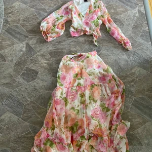 Blommig långkjol och knytblus i pastellfärger - Romantisk långkjol och knytblus med volanger och blommigt mönster i rosa, grön, orange och vit. Kjolen har en luftig och flowy känsla, perfekt för varma dagar. Matcha med en enkel topp eller kör hela setet för en extra söt look. Perfekt till skolavslutningen!