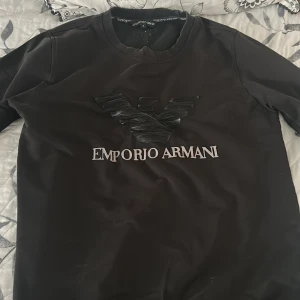 Svart sweatshirt från Emporio Armani - Svart sweatshirt från Emporio Armani med stort 3D-logotyptryck och text på bröstet. Tröjan har rund halsringning, långa ärmar och ribbade muddar vid ärmslut och nederkant. Perfekt för en stilren och trendig look.