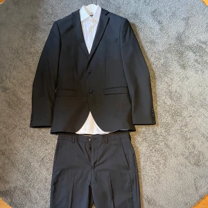 Svart kostym med vit skjorta från Jack & Jones - Klassisk svart kostym med tvåknäppt kavaj, raka kostymbyxor och vit skjorta. Skjortan har knappar framtill och klassisk krage. Har bara använt kostymen en gång till skolavslutning och säljer för den är för liten för mig nu. 