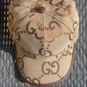 Beige keps från Gucci med GG-mönster - Snygg beige keps från Gucci med det klassiska GG-logomönstret i brunt. Kepsen har böjd skärm, bruna detaljer och justerbart spänne bak i läder. Tillverkad i Italien och gjord i tåligt bomullsmaterial. Perfekt för dig som vill sticka ut med en lyxig accessoar.