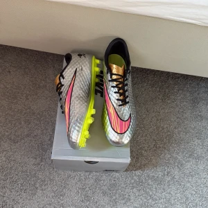 Nike Hypervenom fotbollsskor  - Nike Hypervenom fotbollsskor i metallic silver med guldiga detaljer och neongul sula. Skorna har svarta snören, rosa och gula detaljer på sidan samt svart Nike-logga. Ovandelen har en texturerad yta för bättre bollkontroll. Perfekta för dig som vill sticka ut på planen.
