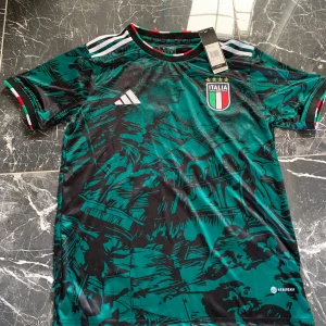 Italien fotbollströja Adidas grön - Snygg Italien fotbollströja från Adidas i grön och svart med unikt grafiskt mönster. Tröjan har korta ärmar, vita Adidas-ränder på axlarna och Italiens klubbmärke på bröstet. Tillverkad i lätt och ventilerande material med Aeroready-teknologi.