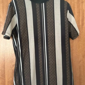 t-shirt  - Snygg t-shirt från River Island i storlek M med vertikala svarta, beige och grå ränder samt diskret mönster i vissa ränder. Klassisk rund halsringning och korta ärmar. Perfekt för dig som gillar grafiska prints och stilren design.