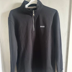 Svart half zip tröja från BOSS - Svart långärmad tröja från BOSS med stilren half zip-dragkedja och broderad logga på bröstet. Tröjan har hög krage och är gjord i mjuk bomull, perfekt för en clean och modern look.