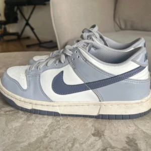 Nike Dunk Low  - Nike Dunk Low sneakers i en clean kombo av ljusblått och vitt skinn med mörkblå swoosh. Klassisk siluett med rund tå, snörning och platt sula. Perfekta för dig som gillar streetstyle och vill ha en fräsch look till vardags.