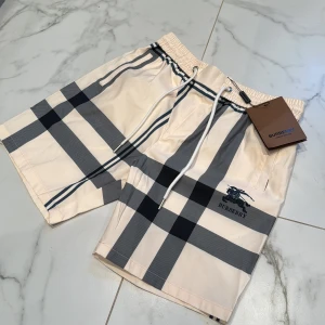 Burberry rutiga shorts med snörning - Snygga shorts från Burberry i klassiskt beige och svart rutigt mönster. De har elastisk midja med vit snörning och broderad Burberry-logga framtill. Tillverkade i lätt polyester, perfekta för sommardagar. Stilsäkra och bekväma med exklusiv känsla.