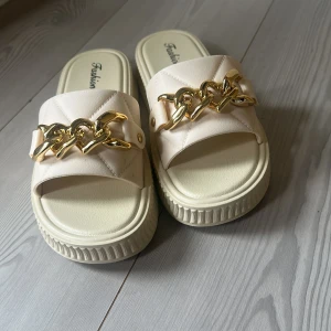 Beige sandaler med guldkedja - Snygga beige sandaler med bred rem i quiltad syntet och chunky guldfärgad kedjedetalj framtill. Platt sula med räfflad kant och öppen tå ger en trendig look. Perfekta för sommardagar när du vill sticka ut lite extra.