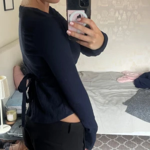 Mörkblå omlottopp med knyt i sidan - Snygg mörkblå omlottopp med v-ringning och långa ärmar. Toppen har knyt i sidan och är lätt croppad, vilket ger en modern och trendig look. Perfekt att styla med både jeans och kostymbyxor. Materialet känns mjukt och följsamt.