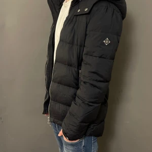 Svart pufferjacka med huva från j lindeberg - Säljer en svart pufferjacka med huva och dragkedja framtill. Jackan har quiltad design, två sidofickor med dragkedja och en liten logga på ärmen. Perfekt för kalla dagar och riktigt skön att bära över en hoodie eller t-shirt. Liten defekt med dragkedjan som kan vara lite trög!